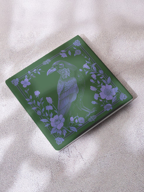 Square ceramic coaster featuring intricate lavender illustration of a raven surrounded by botanical elements on forest green background, with ornate border design and cork backing.